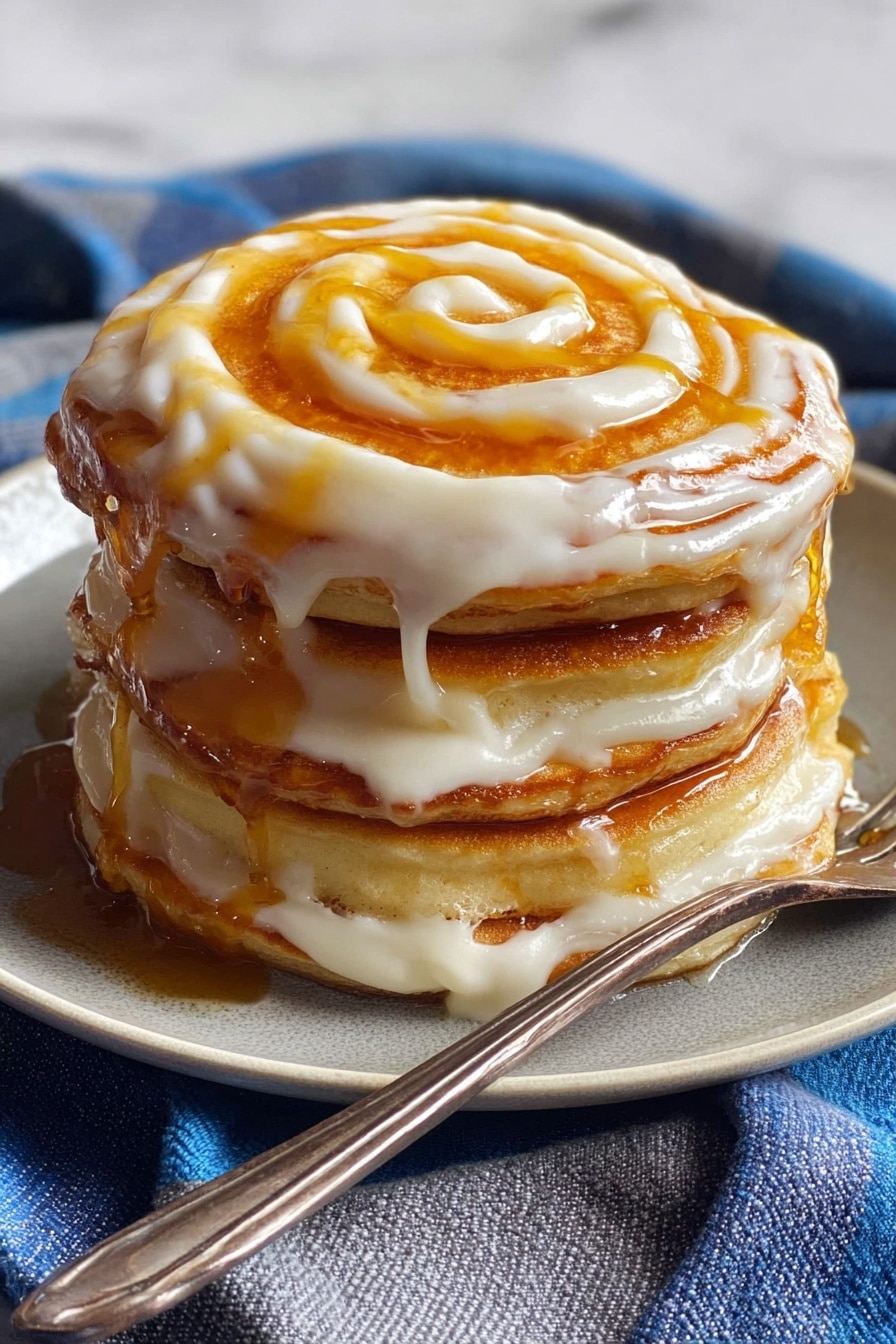 A stack of four thick, golden pancakes sits on a white plate, each layer separated by creamy, smooth off-white icing that drips slightly over the edges, creating a rich texture. On top, there is a spiral of amber-colored syrup swirling over the icing, adding a shiny contrast. A silver fork rests against the plate's edge on a blue and gray striped cloth, and the whole scene is set on a white marbled surface. photo taken with an iphone --ar 2:3 --v 7
