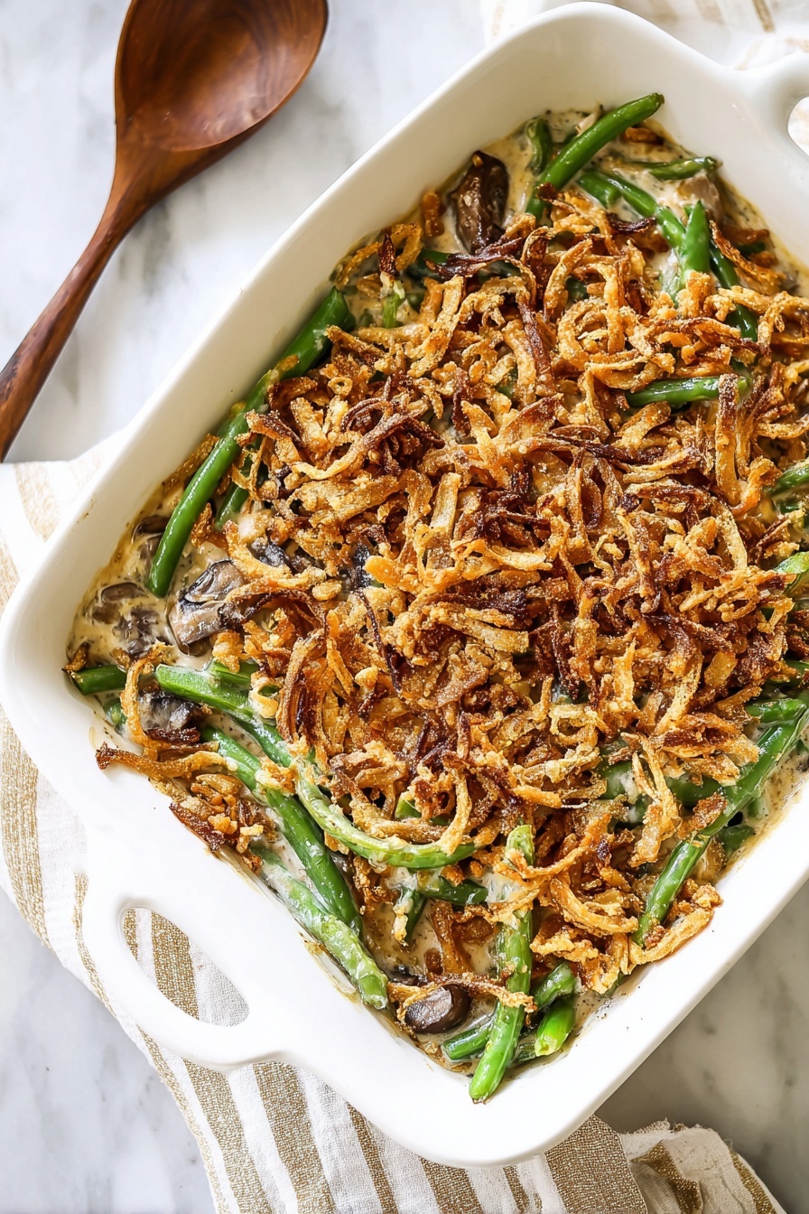 A white baking dish filled with a green bean casserole. The bottom layer has green beans, bright green and slightly shiny. Above the beans is a creamy brownish sauce with small pieces of mushrooms. The top layer is a thick, crispy layer of golden brown and darker fried onion strings spread evenly over the casserole. The dish is placed on a white marbled surface with a striped cream and white cloth underneath. Nearby is a spoon with a wooden handle. Photo taken with an iphone --ar 2:3 --v 7