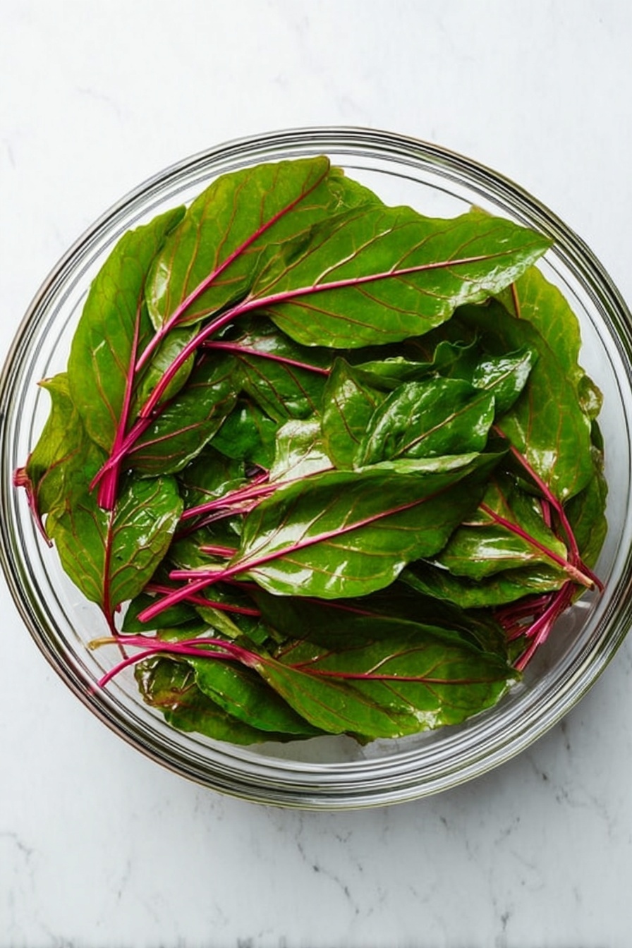 A clear glass bowl holds a large layer of fresh green leaves with red veins and stems, covering the top. The leaves are bright and slightly shiny, spreading evenly across the bowl with some overlapping. The bowl sits on a white marbled surface which adds a clean, simple background to the vibrant green colors of the leaves. photo taken with an iphone --ar 2:3 --v 7