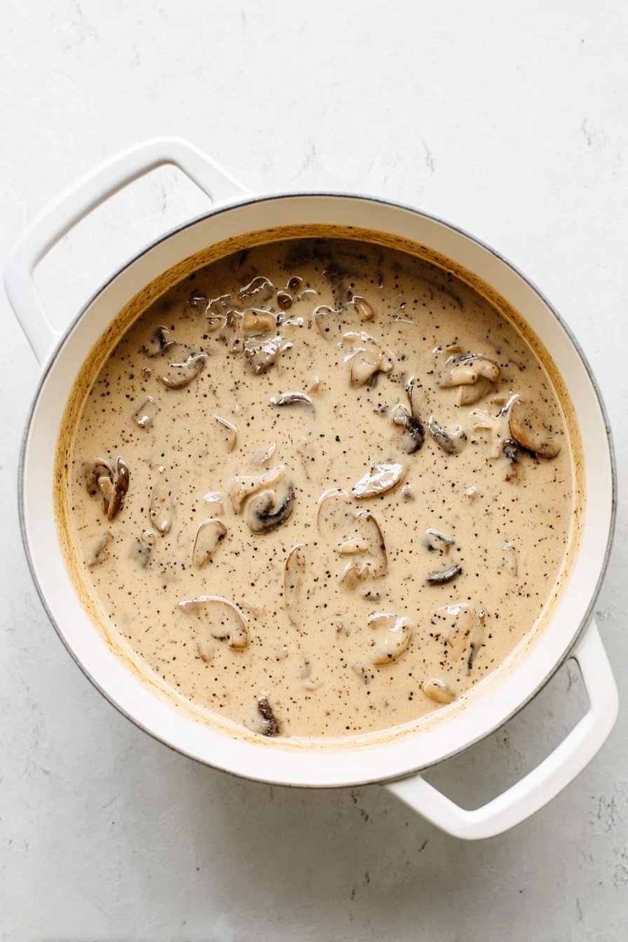 The dish shows a single layer of green beans mixed with a creamy sauce that has small pieces of mushrooms and other vegetables. On top, there is a crispy light brown layer of thin fried onions that adds texture. The food is served in a white bowl with a wooden-handled fork placed on the left side inside the bowl. The background is a white marbled surface with soft natural light. photo taken with an iphone --ar 2:3 --v 7